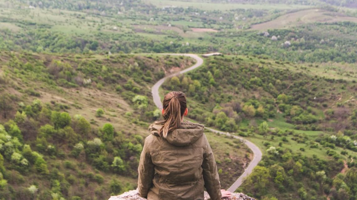 A woman in a jacket sits overlooking a winding road through Romanian countryside in spring.