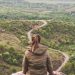 A woman in a jacket sits overlooking a winding road through Romanian countryside in spring.
